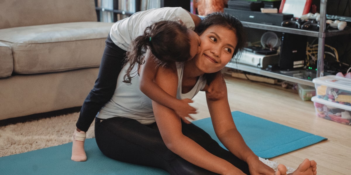 A mother sitting on a yoga mat with her child climbing onto her back