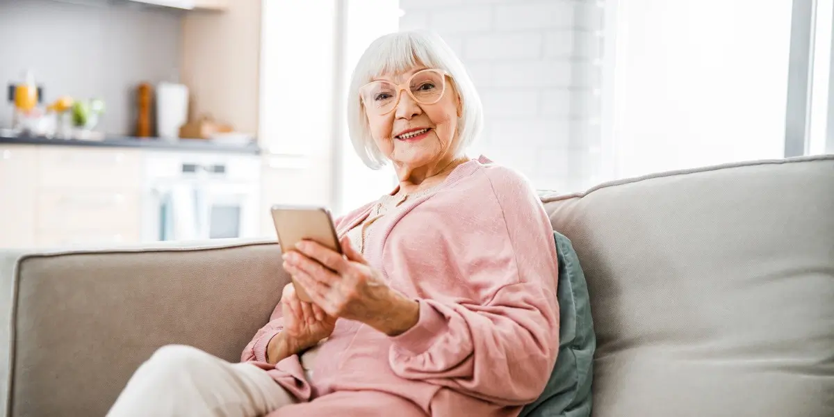 A woman sitting on her couch, holding a mobile phone and smiling at the camera