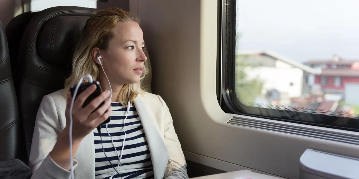 A woman sitting on a train, looking out the window with earphones in, listening to something on her phone