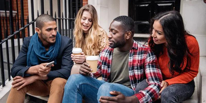 Four people with diverse appearances and backgrounds sitting on some steps, drinking coffee and talking