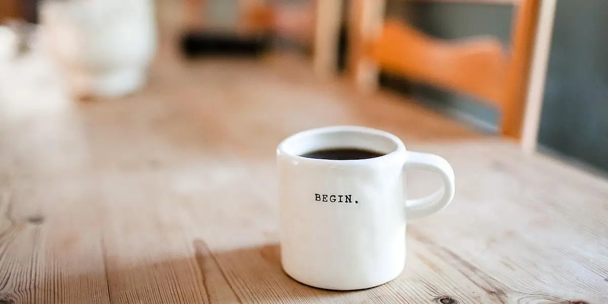 A cup of coffee with the word BEGIN printed on it, sitting on a wooden table