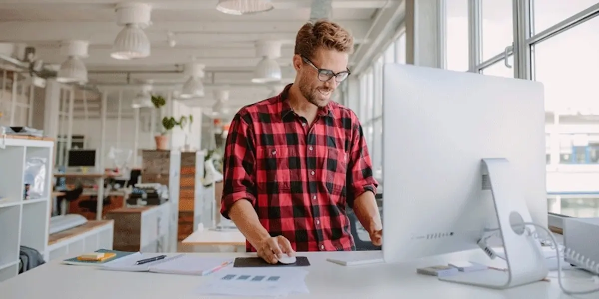 A web designer works at a computer on a standing desk in a bright start-up office.