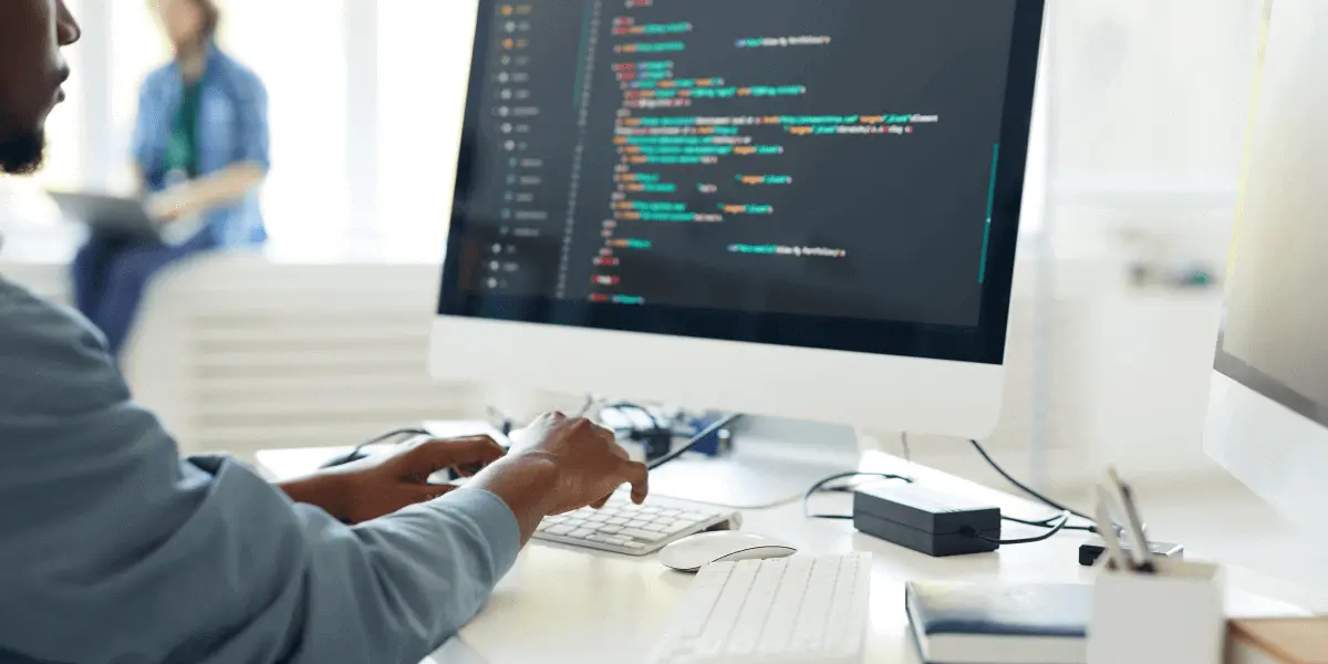 A web developer sitting at a desk in front of a computer, looking at lines of code on the screen.