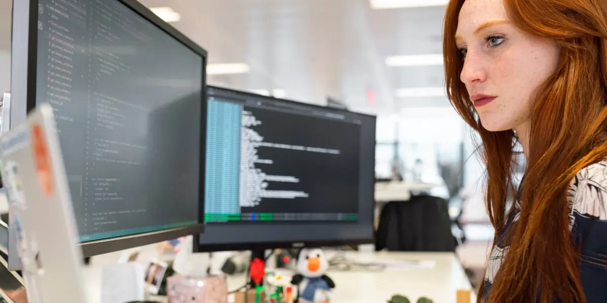 Female web developer sitting at a desk in a startup looking at two screens of code and a laptop