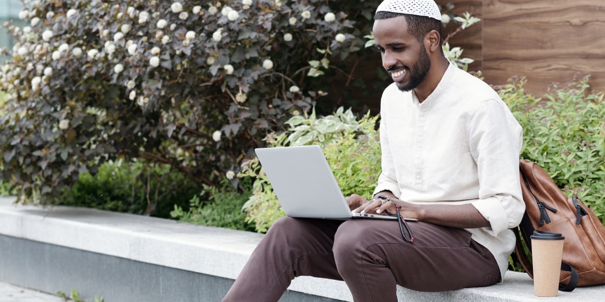 A digital marketing analyst sitting on a wall working on a laptop, with trees and bushes in the background