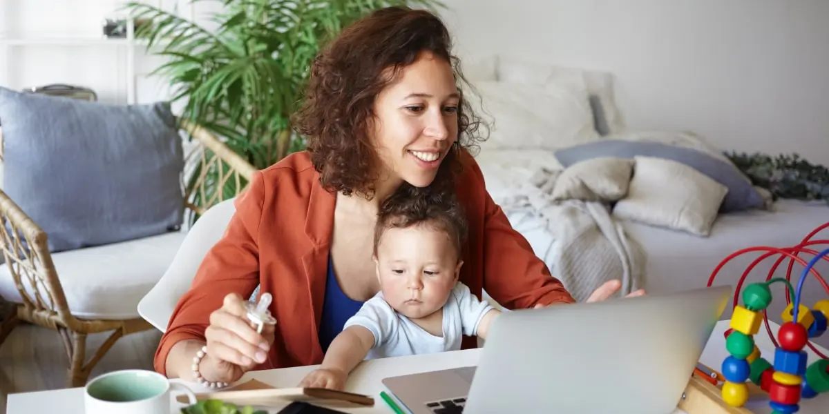 A job applicant sitting at a desk with a baby on their lap, writing their marketing resume on a laptop