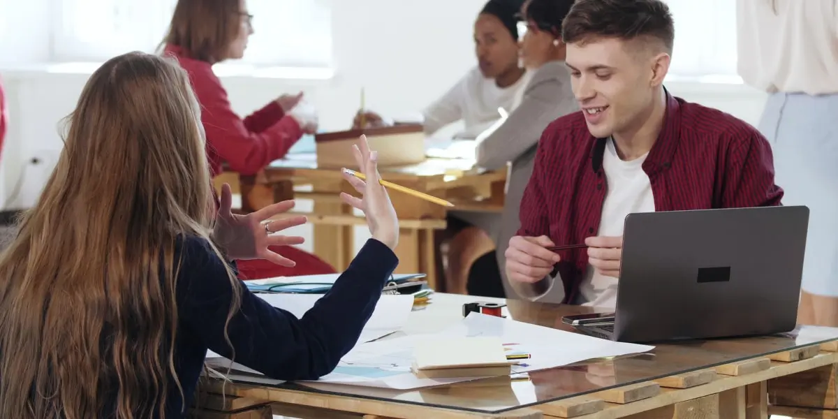 A smiling man and a woman sit at a table during a hackathon, planning their project with a laptop, pens, and paper.