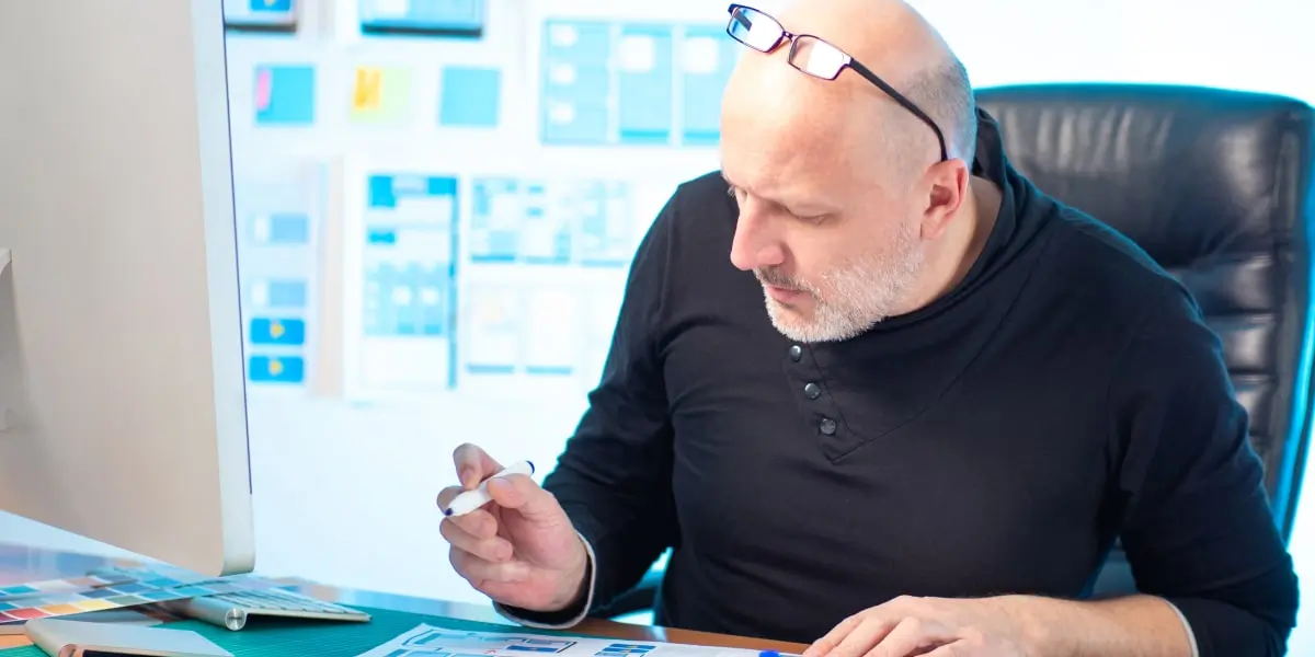 A middle-aged UI designer sits in an office working with color swatches and wireframes.