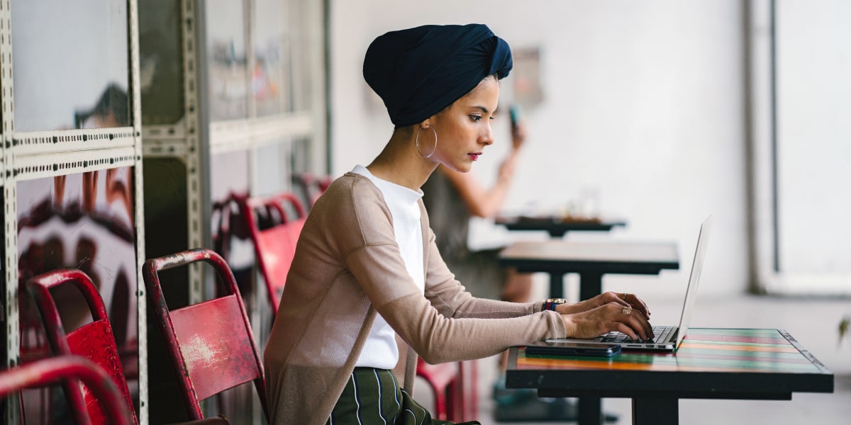 A female designer in a headscarf sits in a startup office working on a laptop.