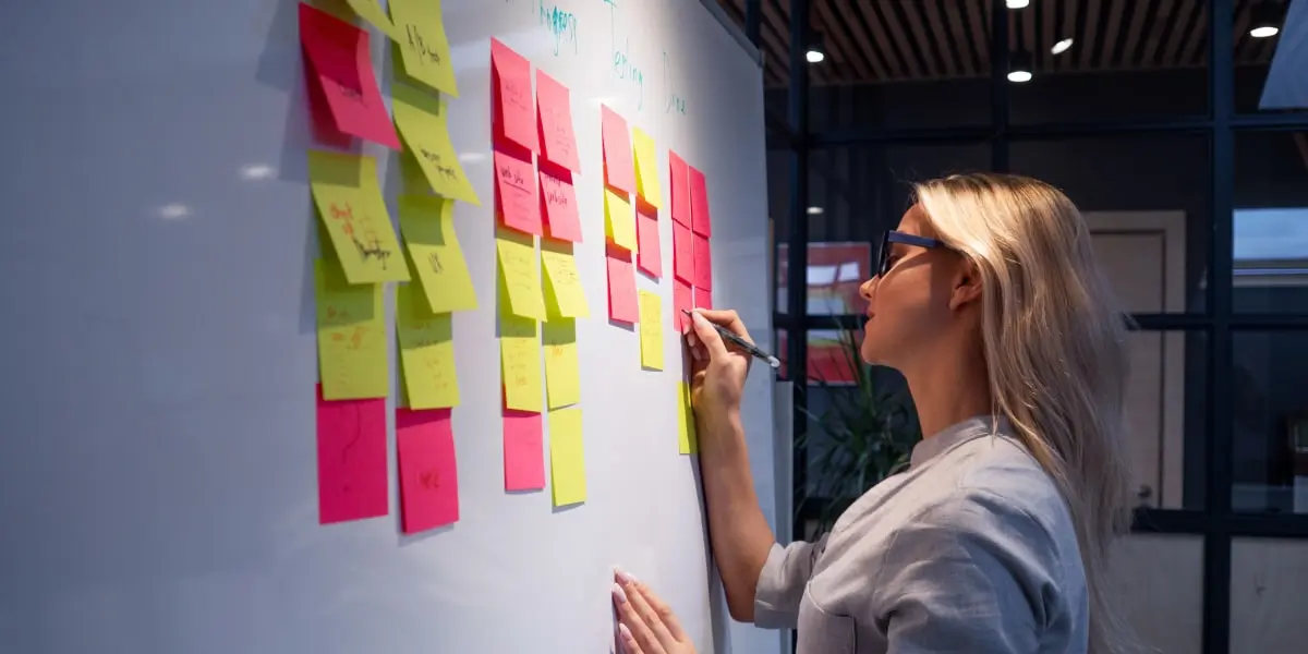 A product manager writes on a sticky note on a whiteboard in an office.