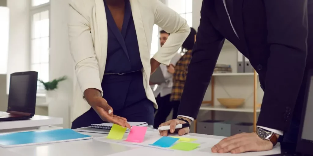 A product manager in an office stands at a table discussing planning with a teammate.