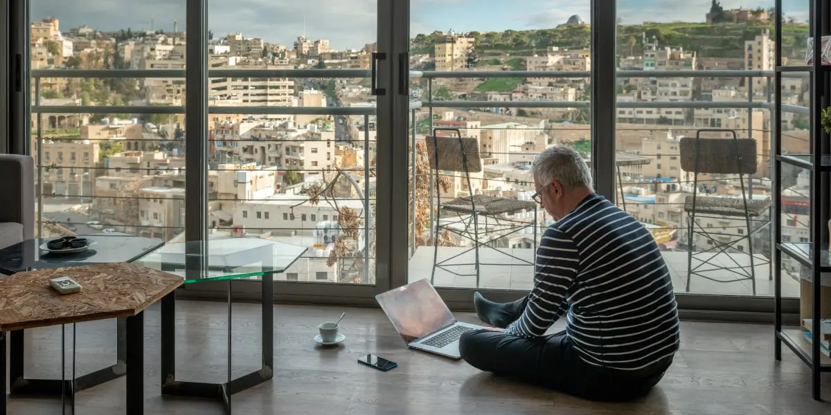 An older product manager sits in an apartment working remotely on his laptop looking over a cityscape.