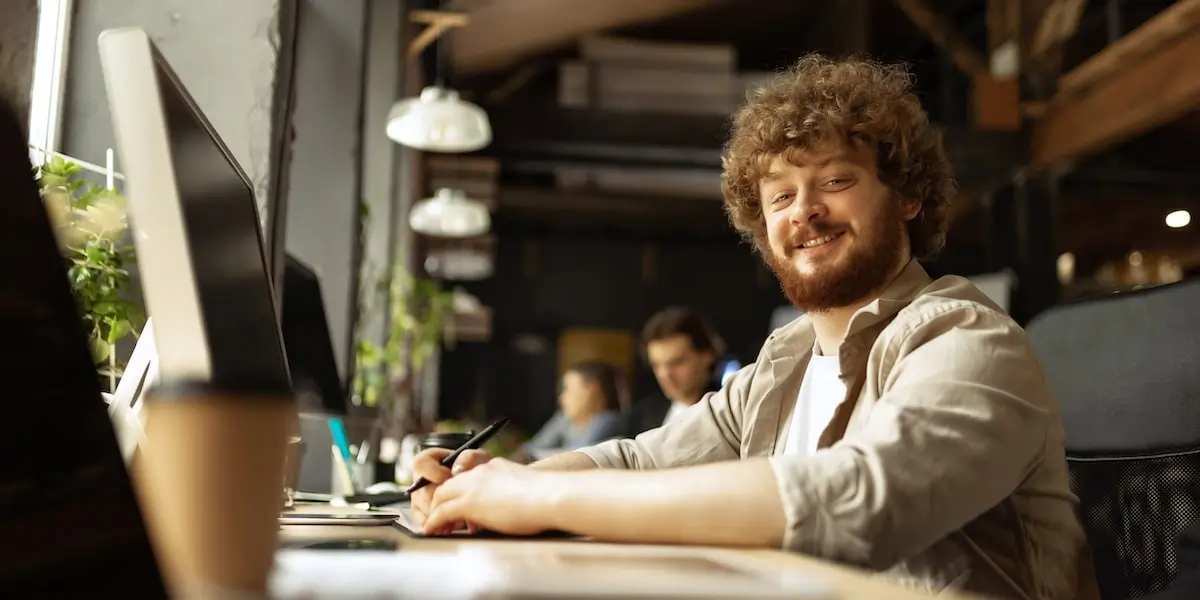 A cloud engineer sits at his computer in a startup office, smiling at the camera.