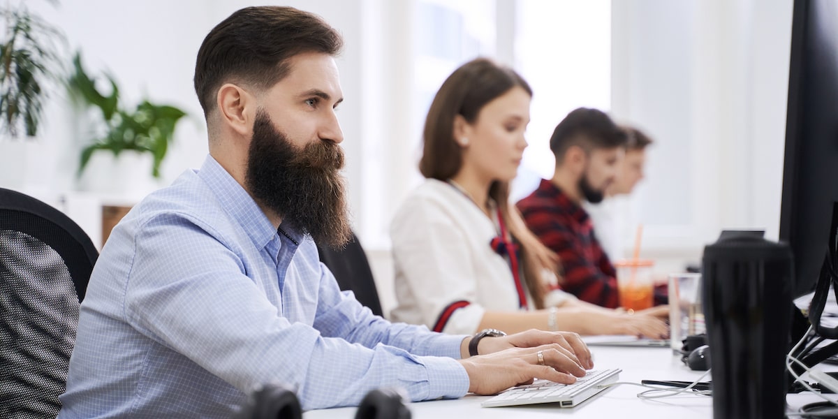 A man sits at a computer in a coding bootcamp with two other classmates behind him.