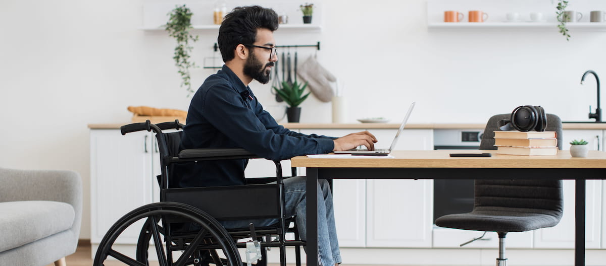 A developer sits in a wheelchair coding on his laptop in his home office.