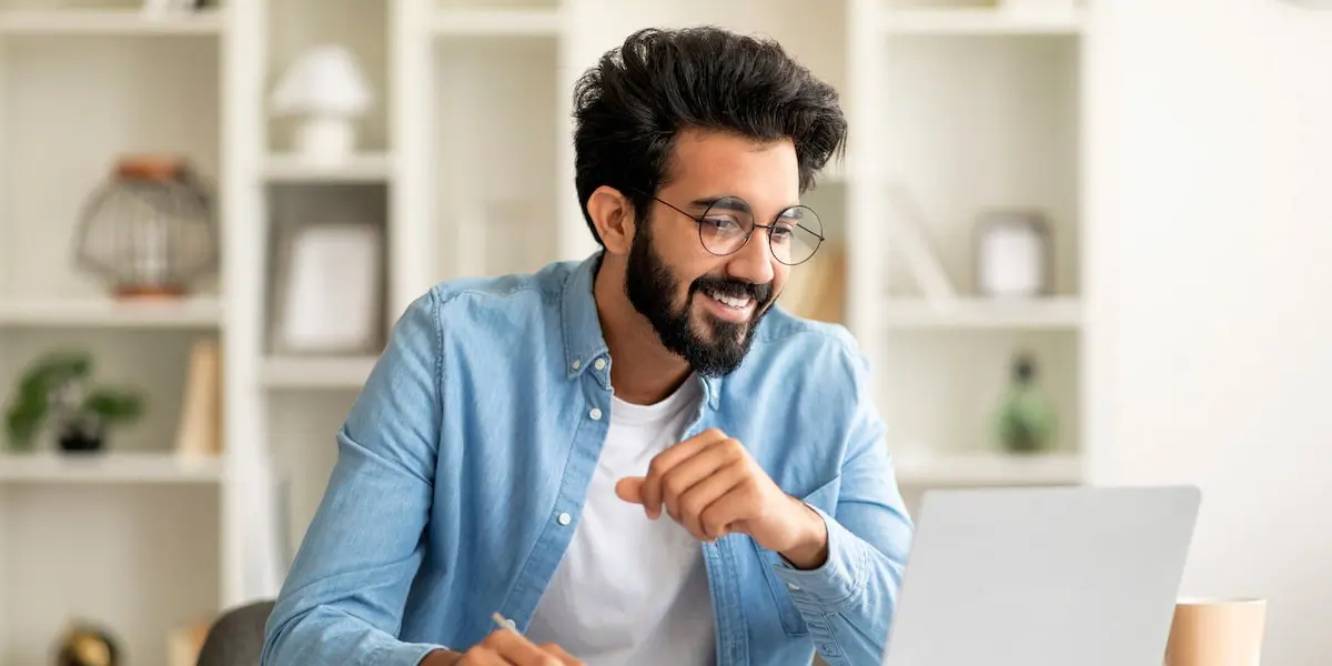 a smiling bearded young adult man holding a pen sits looking for the best online courses for jobs on a laptop
