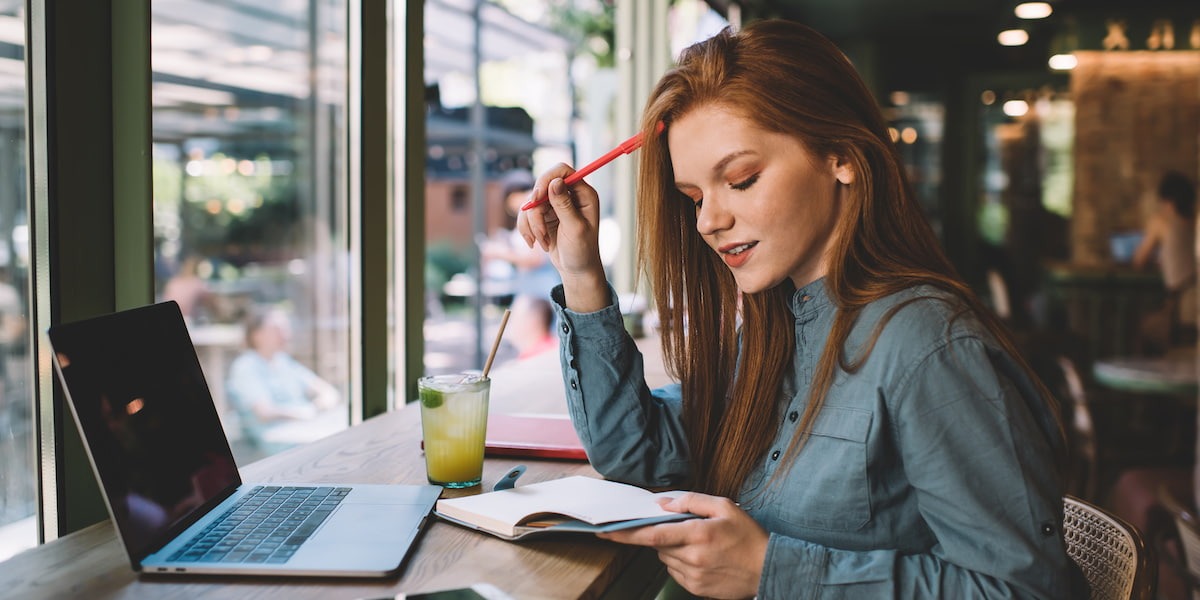 A young redhead sits in a coffee shop at the window with a notepad and computer looking up the best product design courses