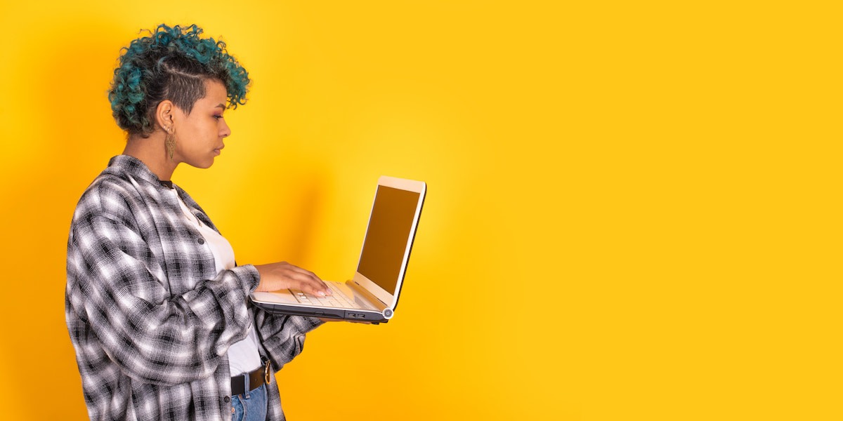 A side profile of a young woman looking at her laptop against a yellow backdrop