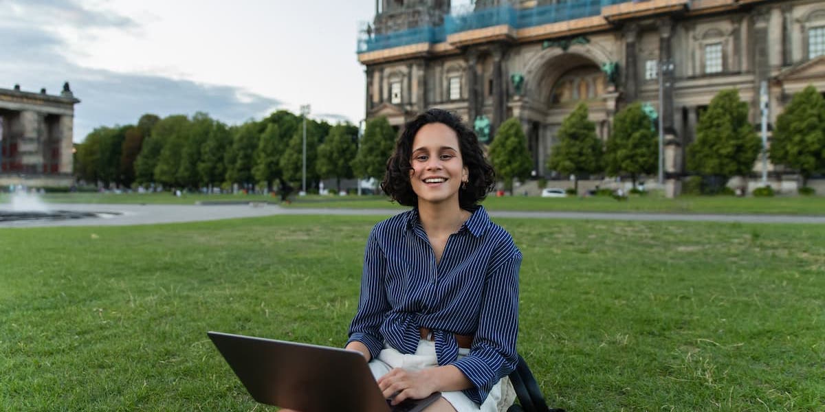 A woman smiles with her laptop as she sits on the lawn in front of the Berliner Dom in Berlin, working at her Germany tech job.