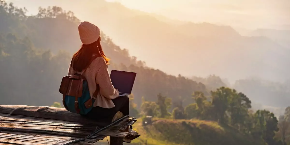 A woman wearing a beanie sits with her laptop on a scenic lookout point.