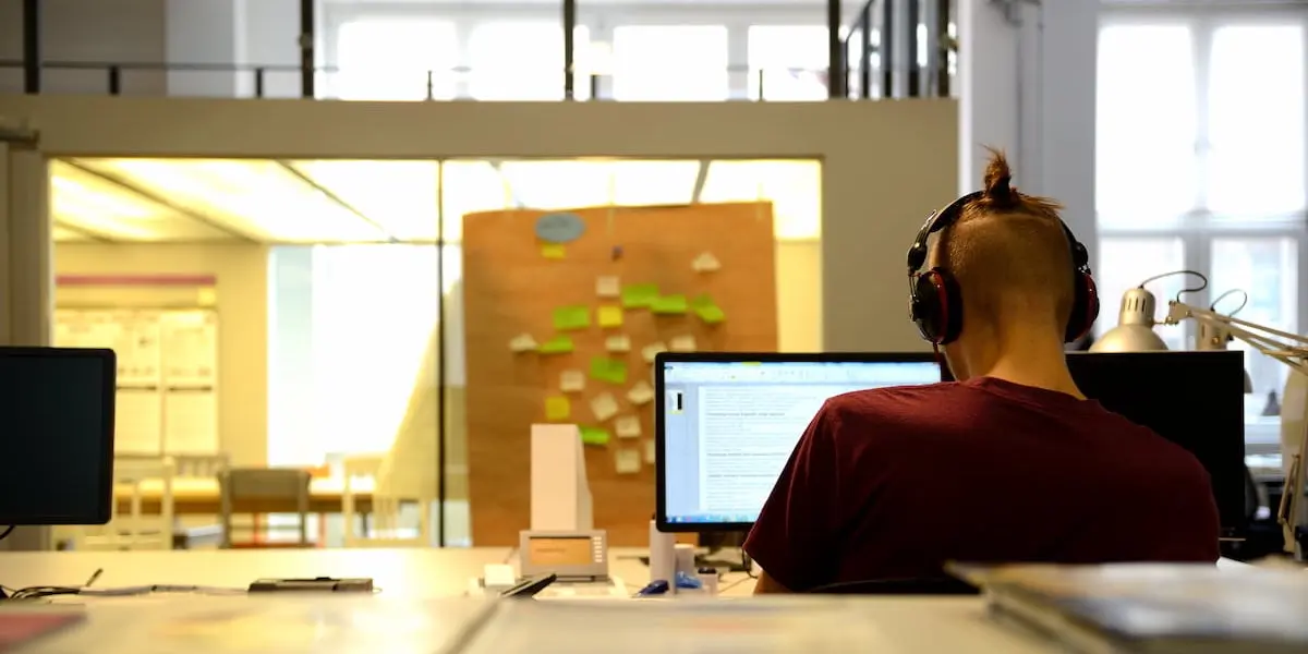 A UX designer in Berlin sits at his computer with headphones and a corkboard of postits in the distance.