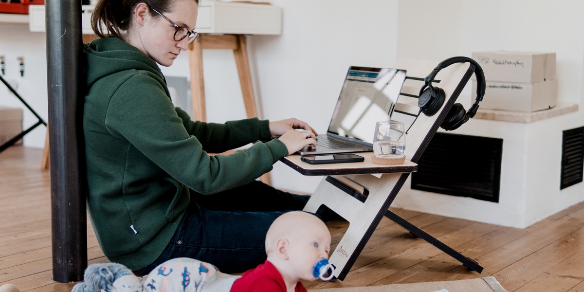 A woman sitting at a makeshift desk on the floor, working at a laptop with her infant nearby