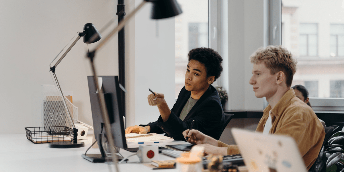 Two male web developers sitting at a table in a bright startup office advising each other on code