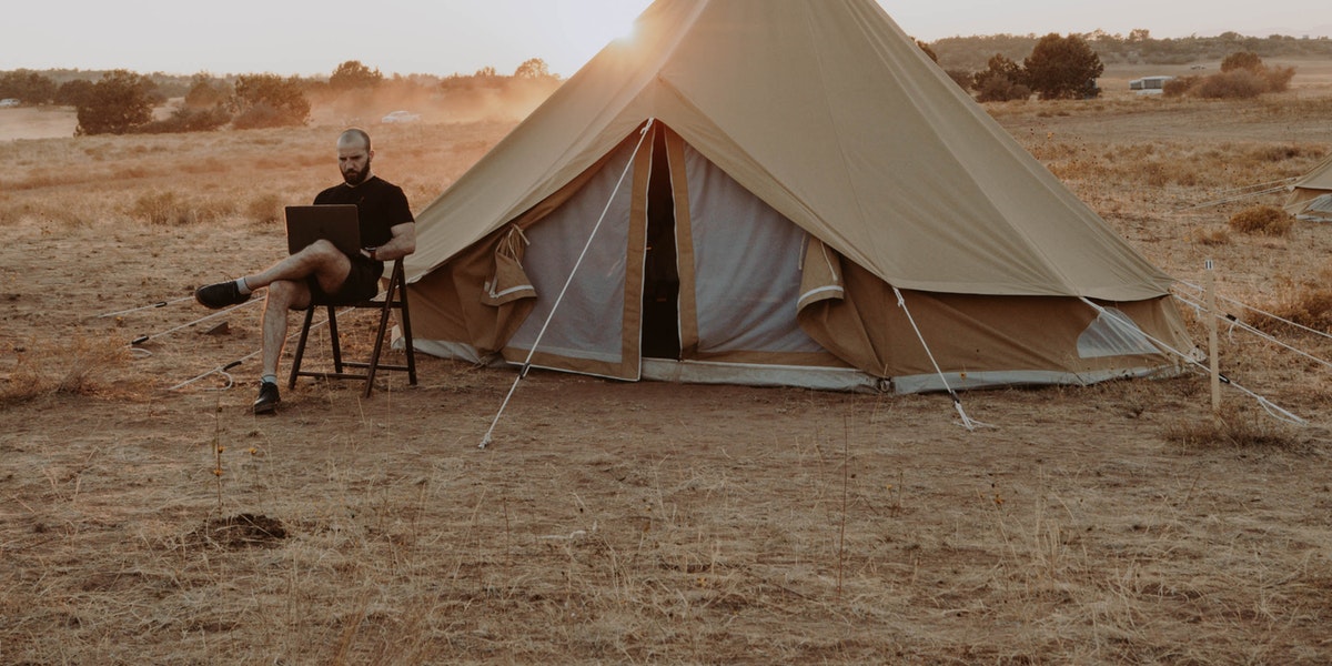 Man sits in a desert beside a tent sitting with a laptop working on code.